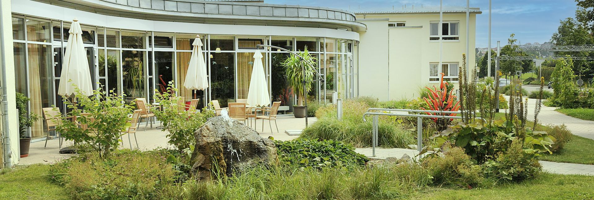 Geschwungene Glasfassade mit Terrasse, Sonnenschirmen, kleinem Springbrunnen und Garten