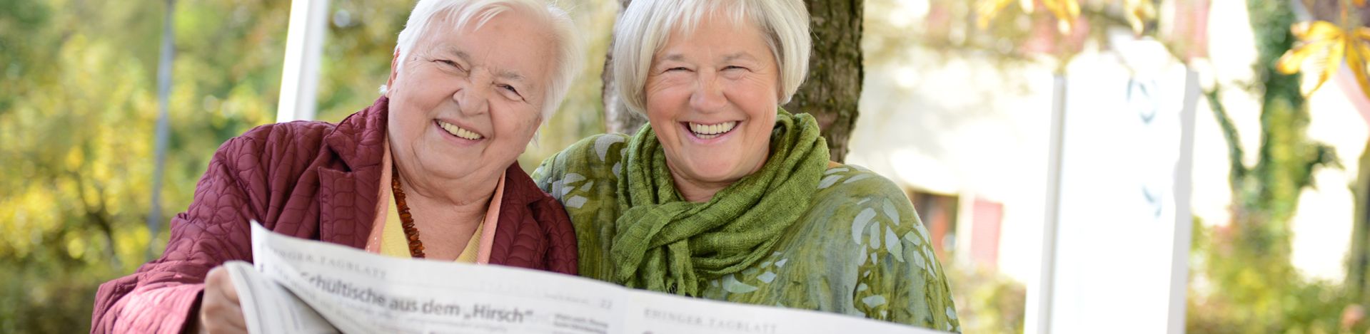 Zwei ältere Frauen lächeln auf einer Parkbank und lesen Zeitung